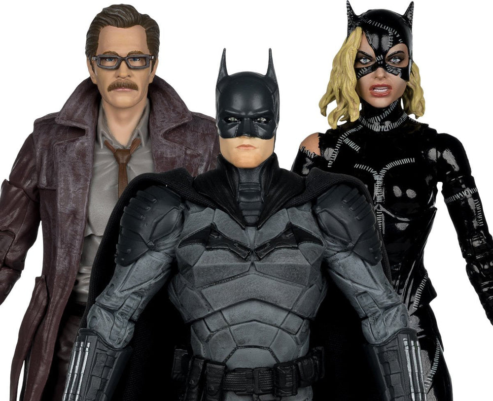 Three Batman-themed collectible figures posed together against a plain white background: a central Batman in a dark armored suit with cape, a woman in a black cat-ear mask and costume on the right, and a man in a brown trench coat and glasses on the left.